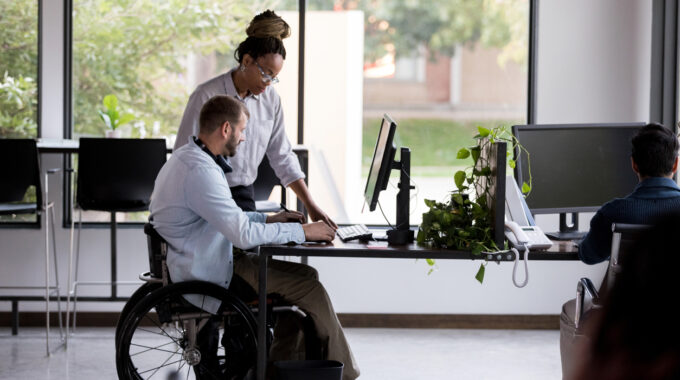 Diversity, Equity & Inclusion At Work; Mid Adult Wheelchair-bound Businessman And A Female Colleague Review A Current Project. They Are Looking At Something On A Desktop Computer.